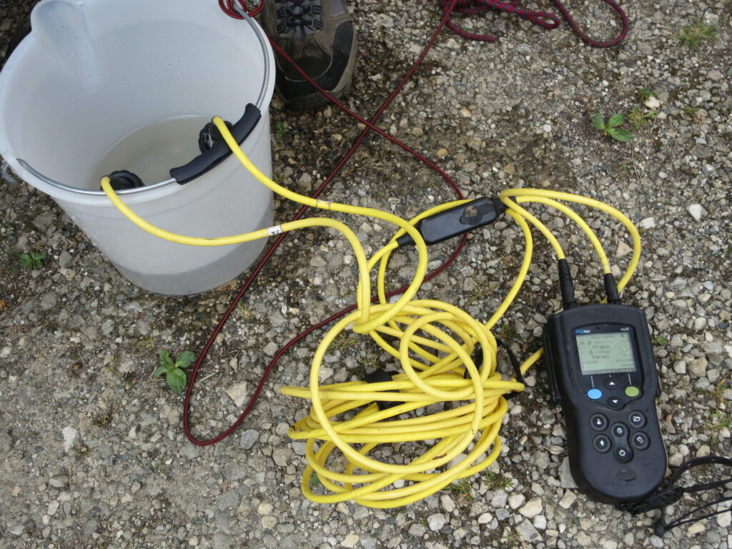 The image displays a multimeter device measuring parameters of a water sample in a bucket.